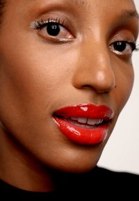 Klarröd, glänsande läppstift på en jämn, medelhudton. Närbild av läpparna och en del av ansiktet med väldefinierade ögonfransar och subtil ögonmakeup.