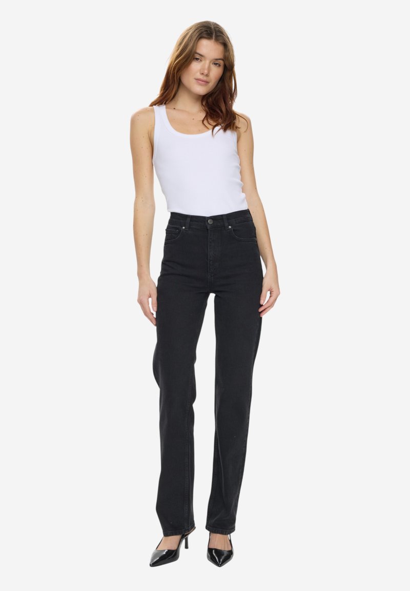 Femme à la peau claire et aux cheveux bruns portant un haut blanc sans manches, un jean noir taille haute et des talons pointus noirs, debout de manière détendue.