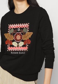 Sudadera negra con intrincados bordados multicolores de hojas y flores, con la palabra "WANDERER" arriba y "Maison Scotch" debajo.