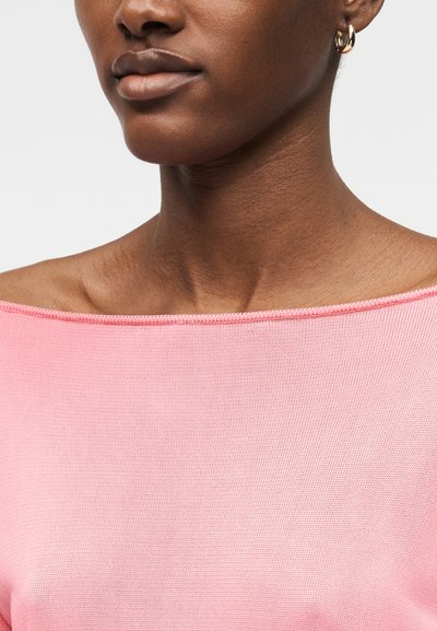 Γυναίκα που φοράει ροζ τοπ με ανοιχτούς ώμους και μικρό χρυσό κρίκο στο αυτί, εμφανίζοντας λεπτομέρειες του λαιμού και του κάτω μέρους του προσώπου σε λευκό φόντο.