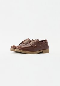Pantofi din piele maro cu vârf rotunjit, două deschideri pentru șireturi, gauri aurii pentru ochi și o talpă din cauciuc texturat. Design minimalist, stil casual.