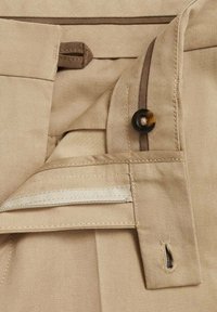 Pantaloni beige con zip, bottone marrone e chiusura a gancio. Il tessuto ha una texture liscia con dettagli di cucitura discreti.