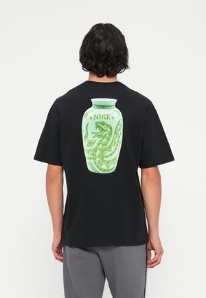 Oseba s kratkimi temnimi lasmi, oblečena v črno majico z zeleno kačo in napisom "NIKE" na hrbtu, v kombinaciji s sivimi hlačami.