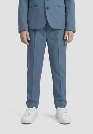 Niño con pantalones azules a medida y blazer a juego con zapatillas blancas, de pie frente a un fondo claro y sencillo.