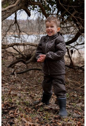 Braune gesteppte Jacke und Hose, graue Gummistiefel. Kind steht in einem Waldgebiet, umgeben von getrockneten Blättern und Zweigen.