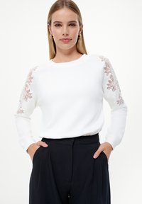 Sudadera blanca con bordado floral en rosa pálido en los hombros, de mangas largas y cuello redondo, combinada con pantalones negros.