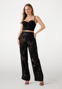 Čierny kvetinový čipkový crop top so štruktúrovaným výstrihom a tenkými ramienkami, doplnený o priehľadné čierne nohavice s širokými nohavicami a pevným lemom.