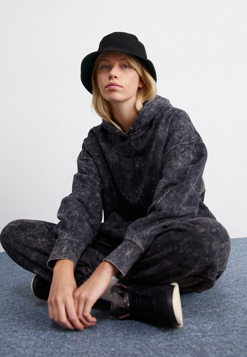 Sudadera negra con diseño de tie-dye y pantalones de chándal a juego de corte holgado. Llevado con un sombrero tipo cubo negro y zapatillas negras sobre una alfombra azul.
