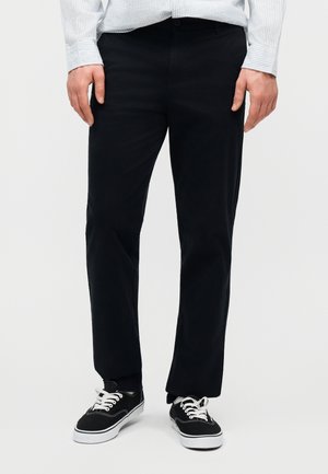 Άνδρας που φοράει μαύρο παντελόνι ίσιας γραμμής, μαύρα χαμηλά αθλητικά παπούτσια με λευκά κορδόνια και ανοιχτό πουκάμισο με μακριά μανίκια και ρίγες.