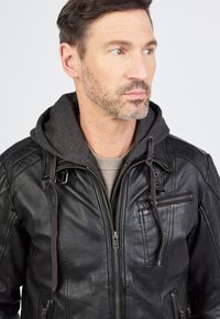 Zwarte leren jas met een grijze hoodie voering. Kenmerken zijn onder andere ritsen, zakken en een gestructureerde afwerking. Getailleerde pasvorm met stikseldetails.