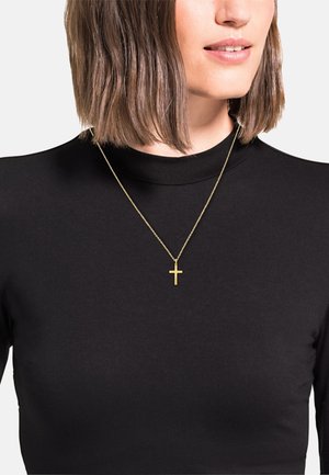 Złoty wisior w kształcie krzyża na cienkim łańcuszku, noszony na czarnym golfie. Krzyż ma minimalistyczny i płaski design.