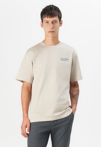 Beige Baumwoll-T-Shirt mit kurzen Ärmeln und Rundhalsausschnitt. Mit blauer Schrift "SOHO Social Coffee CLUB" auf der linken Brustseite.
