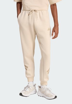 Mann trägt beige Jogger-Hosen mit Kordelzug in der Taille und Logodetails, kombiniert mit weißen und beigen Sportschuhen und weißen Socken.