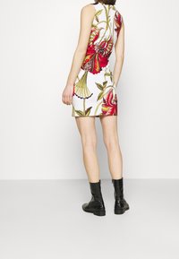 Robe sans manches à imprimé floral en blanc, présentant des motifs vifs rouges et jaunes. Le mannequin porte des bottines noires à la cheville. Vue de dos affichée.