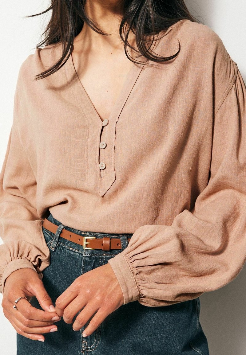 Blouse rose blush à col en V avec trois boutons, manches longues bouffantes et poignets froncés, associée à une ceinture en cuir marron et un jean en denim.