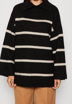 Zwarte trui met brede crèmekleurige horizontale strepen, lange mouwen en een losse kraag, gedragen met camelkleurige broek.