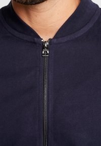 Zbliżenie na szyję i dolną część twarzy mężczyzny, który ma na sobie ciemnogranatowy sweter rozpinany na zamek błyskawiczny, z metalowym zamkiem podciągniętym do góry.