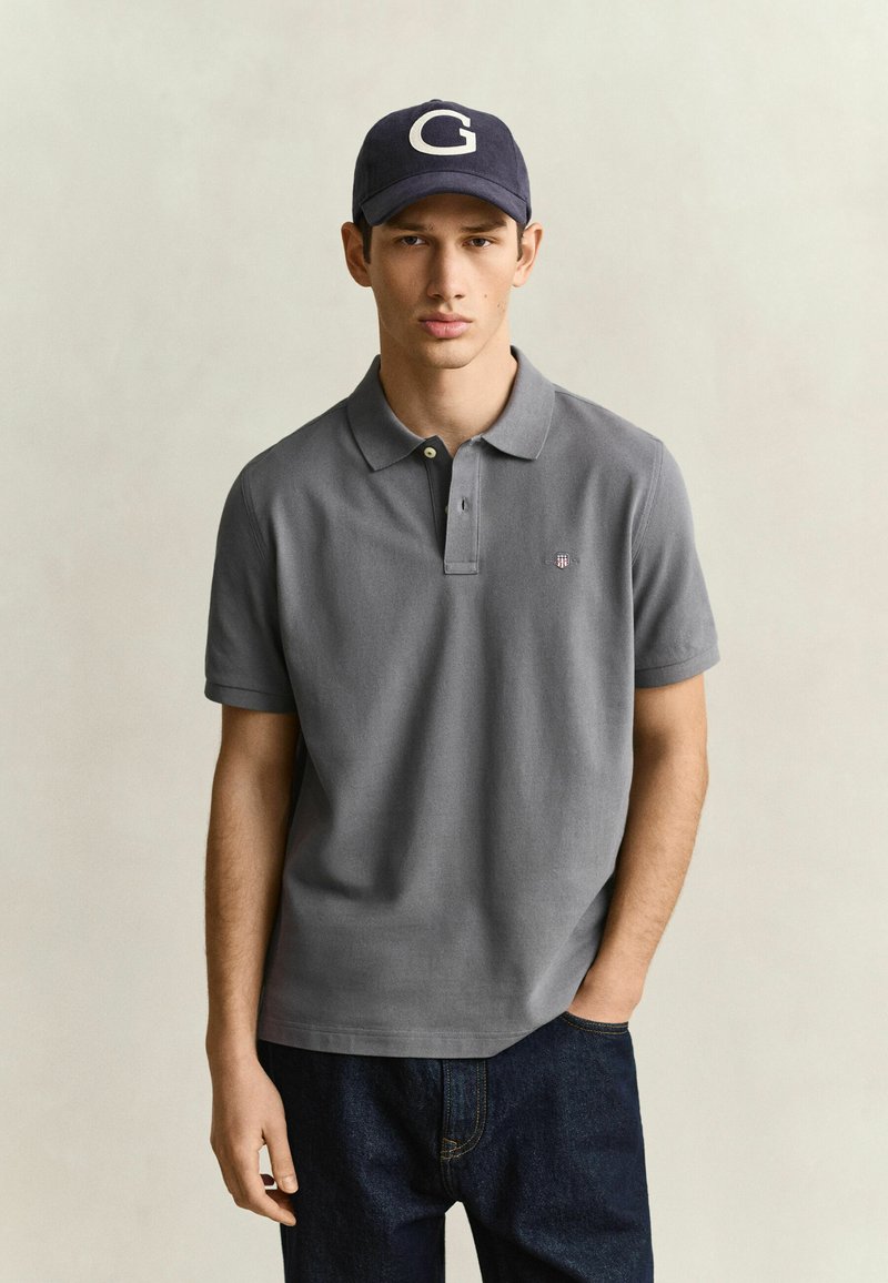 Jeune homme portant un polo gris, un jean foncé et une casquette bleu marine avec une lettre G blanche, debout avec une main dans la poche.