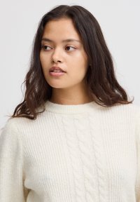 Pull crème avec une texture côtelée et un motif de côtes central. Encolure haute avec une coupe décontractée et un matériau doux.
