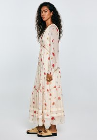 Lange, vloeiende crèmekleurige jurk met een bloemenprint in rood en blauw, kantdetails bij de taille en lange mouwen. Draag met tan kleurige sandalen.