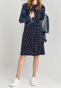 Marinblå klänning med blåa cirkelmönster, korta ärmar och midjeband, bärs under en denimjacka och matchas med vita sneakers.