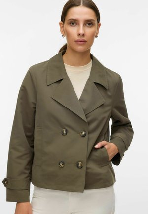 Naine, kes kannab oliivrohelist topeltlõikelist jopet laiade kraedega ja beeži särki, käsi taskus, neutraalse ilmega, valgel taustal.