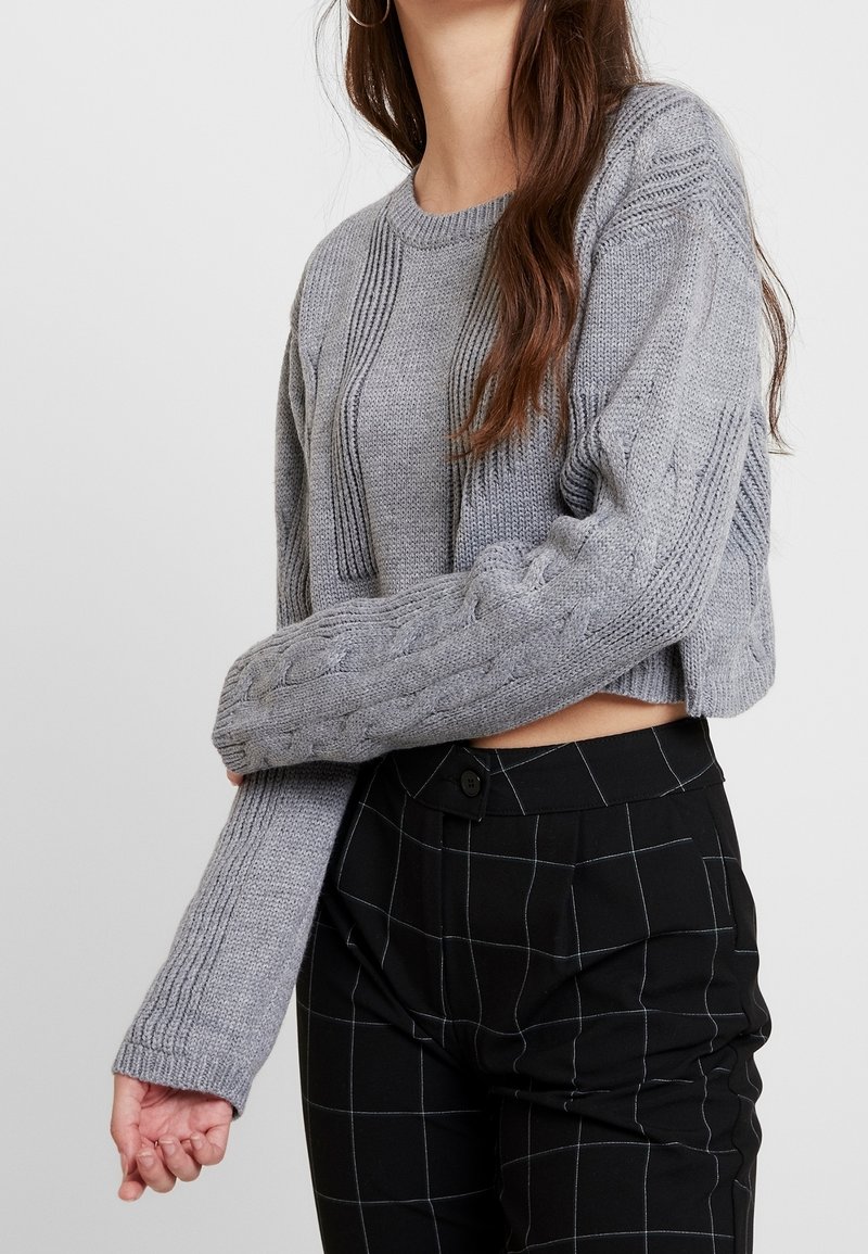 Szary dziergany krótki sweter z prążkowanymi i warkoczowymi wzorami. Długie rękawy, lekko oversize'owy krój. Połączony z czarnymi spodniami w kratę.
