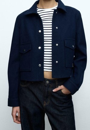 Person trägt eine marineblaue kurze Jacke mit silbernen Knöpfen, ein schwarz-weiß gestreiftes Crop-Top und dunkelblaue Jeans, eine Hand in der Tasche.