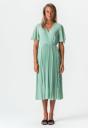 Egy nő szemben áll világoszöld, redős midi ruhában, flutter ujjakkal és fekete, nyitott orrú szandálban, fehér háttér előtt.