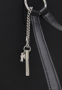 Sac à main noir texturé avec une chaîne en argent et un porte-clés arborant la marque Lacoste, incluant un petit charm en forme de crocodile.