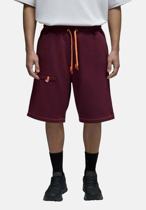 Homme portant un short bordeaux avec des cordons orange et un détail de poche, chaussettes noires et baskets noires, debout sur fond blanc.