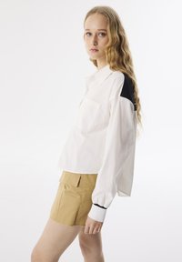 Camicia bianca a maniche lunghe con una toppa nera sulla spalla e una tasca sul petto, abbinata a pantaloni corti beige con due tasche frontali e orli sfrangiati.