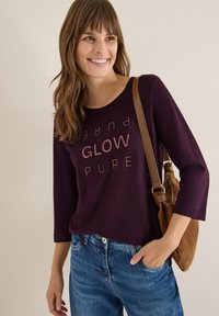 Maglietta a maniche lunghe viola con testo "GLOW PURE" in rosa chiaro, indossata con jeans blu e una borsa a tracolla marrone. Tessuto morbido, vestibilità rilassata.