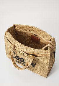 Sac fourre-tout en paille tissée avec lettrage noir, couleur beige, deux poignées de transport, et une fermeture zippée, présentant une doublure intérieure lisse.