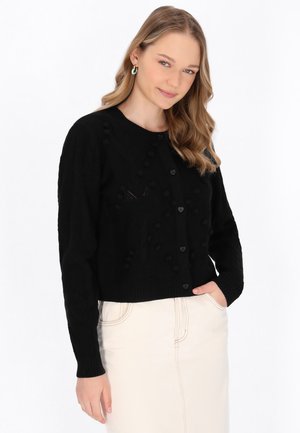 Frau mit welligem blondem Haar, die eine schwarze, geknöpfte Strickjacke und einen hellen Rock trägt, lächelt leicht mit einer Hand in der Tasche.