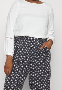 Maglietta bianca a maniche lunghe con tessuto testurizzato e dettagli in pizzo, abbinata a pantaloni elasticizzati bianchi e neri con motivo e tasche laterali.