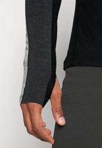 Långärmad atletisk topp i svart och grått texturerat tyg, med sidopaneldetalj och tumhål vid muddarna.