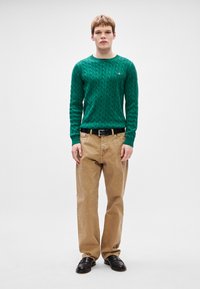 Πράσινο πουλόβερ με πλέξη, μπεζ παντελόνι, μαύρη ζώνη και μαύρα παπούτσια. Το πουλόβερ διαθέτει υφαντό μοτίβο και ριμπ μανσέτες.