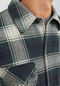 Cămașă din flanel cu un model în carouri verzi și albe, guler butonat, nasturi negri și un buzunar pe piept cu o textură moale.
