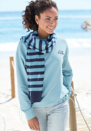 Young woman wearing light blue sweatshirt and striped scarf standing on wooden pathway by the beach, smiling and looking to the side.