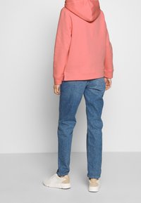 Haut à capuche rose, associé à un jean en denim bleu clair et des baskets blanches avec des accents beige. Accent sur la coupe décontractée et le tissu doux.