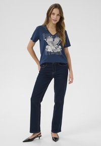 T-shirt navy a collo V con grafica di un'aquila bianca e testo "FOREVER STRONG", abbinato a jeans neri a gamba dritta e décolleté neri con cinturino.