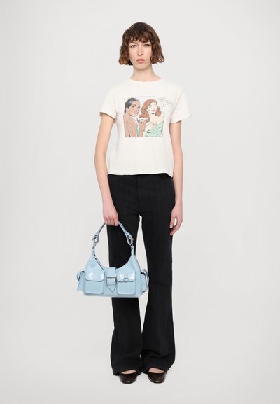 Sac à main en cuir verni bleu clair avec deux poches avant et une bandoulière robuste. Le modèle porte un t-shirt à motifs et un pantalon noir à rayures.