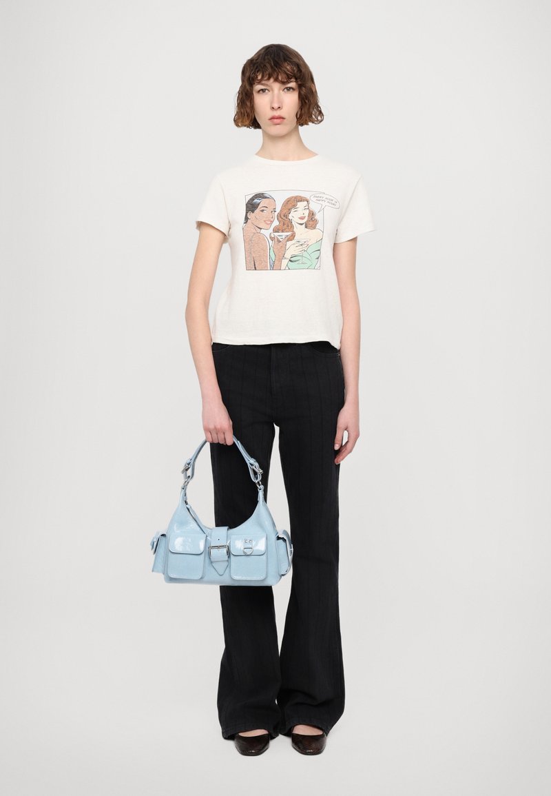 Borsa a tracolla in pelle verniciata blu chiaro con due tasche frontali e una tracolla robusta. Il modello indossa una maglietta grafica e pantaloni neri a righe sottili.