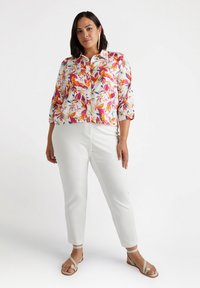 Top a manica corta a fiori con base bianca, caratterizzato da vivaci motivi arancioni, rosa e viola; abbinato a pantaloni bianchi aderenti e sandali beige.