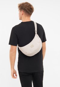 Borsa a tracolla in tessuto beige con una texture liscia, forma curvata e una tracolla regolabile. Presenta un piccolo marchio sul davanti.
