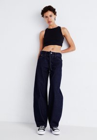 Juoda trumpa tankų palaidinė derinama su plačiomis tamsiomis džinsinėmis kelnėmis, turinčiomis raištelį ir subtilų siuvinėjimą. Juodi ir balti sportbačiai užbaigia išvaizdą.