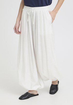 Kobieta nosząca luźne białe spodnie w cienkie paski z elastyczną talią oraz czarne wsuwane buty, stojąca na tle jednolitym.