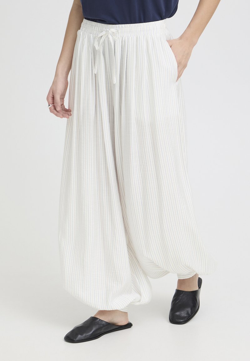 Žena stojaca na rovnom pozadí, oblečená v voľných bielych prúžkovaných nohaviciach s elastickým pásom a čiernych slip-on topánkach.