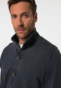 Dark denim jacket with a high collar, featuring two large brown buttons, visible stitching details, and a textured fabric.